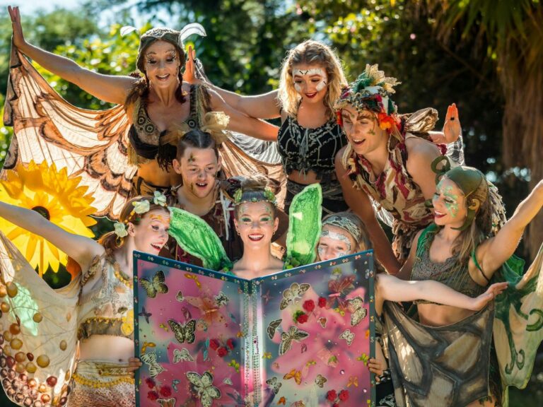 Seven people dressed as fairies gather around a beautifully decorated book in the gardens.